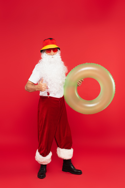 Santa Claus ve slunečních brýlích drží plavecký prsten a ukazuje jako na červeném pozadí - Fotografie, Obrázek