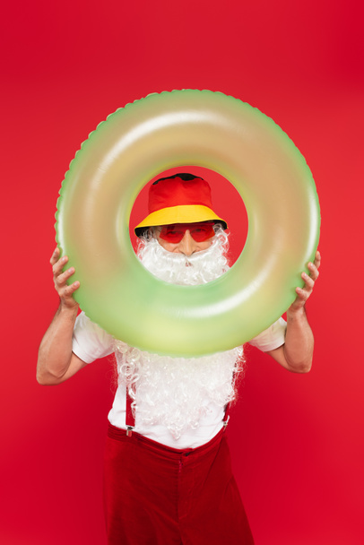 Santa Claus en panama y gafas de sol mirando a la cámara a través de un anillo de natación aislado en rojo  - Foto, Imagen