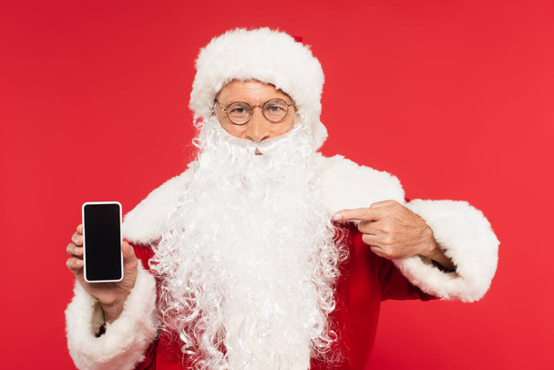 Santa Claus apuntando a teléfono inteligente con pantalla en blanco aislado en rojo  - Foto, imagen