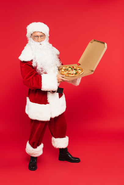 Santa Claus en traje y anteojos sosteniendo pizza en caja sobre fondo rojo - Foto, imagen