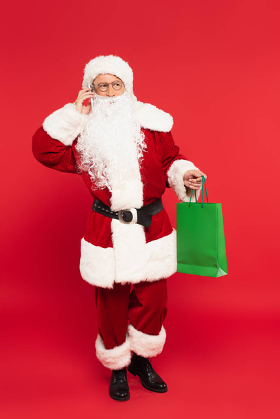 サンタさんがスマートフォンで話して、赤い背景にショッピングバッグを持っている - 写真・画像