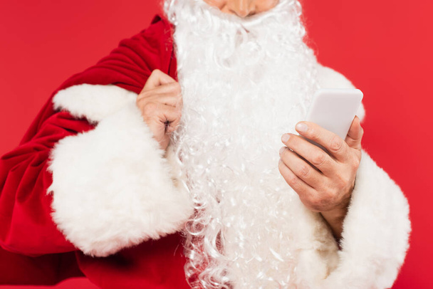Cropped view of smartphone in hand of blurred santa claus with sack isolated on red  - Foto, afbeelding