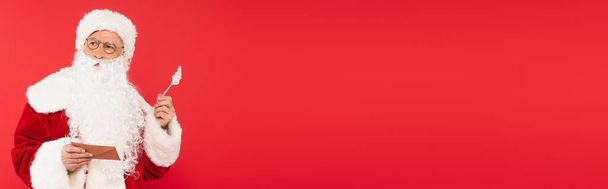 Pensive santa claus tenant enveloppe et stylo isolé sur rouge, bannière  - Photo, image