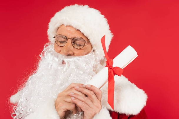 Santa Claus se zavřenýma očima drží papír s stuhou izolovanou na červené  - Fotografie, Obrázek
