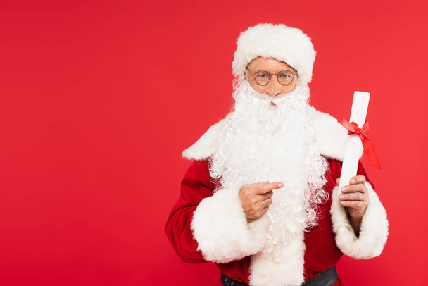 Père Noël pointant vers le papier avec arc isolé sur rouge  - Photo, image