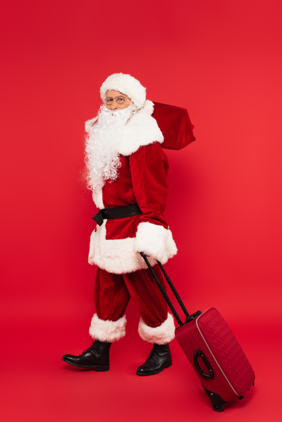 Санта-Клаус в костюме держит чемодан и мешок на красном фоне - Фото, изображение