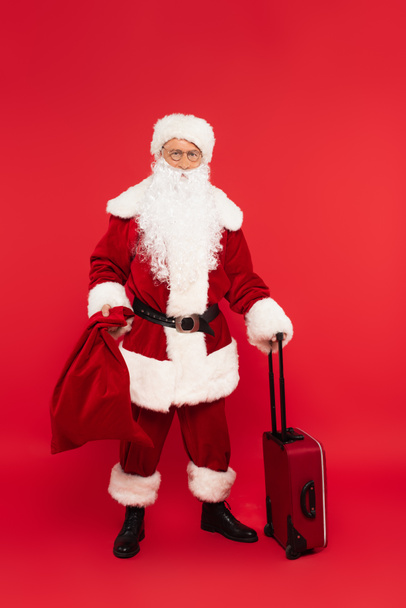 Kerstman bedrijf zak en koffer op rode achtergrond - Foto, afbeelding