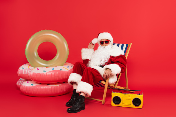 Santa claus in sunglasses sitting on deck chair near boombox and swim rings on red background - Photo, Image