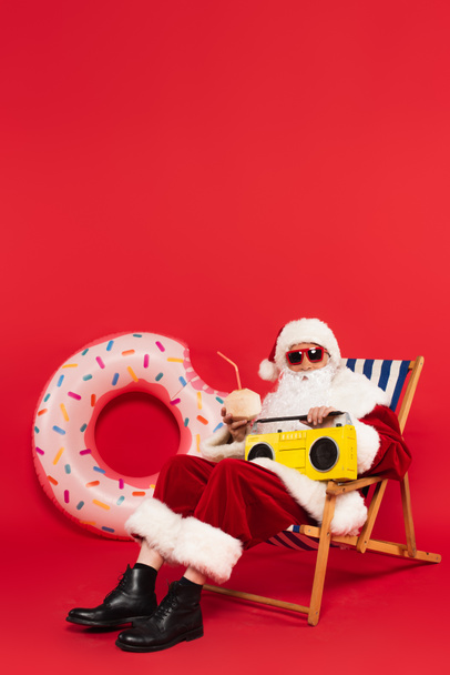 Kerstman in zonnebril met retro boombox en cocktail in kokosnoot op ligstoel nabij zwemring op rode achtergrond - Foto, afbeelding