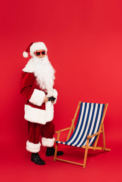 Санта-Клаус в костюмах и солнцезащитных очках стоит возле шезлонга на красном фоне - Фото, изображение