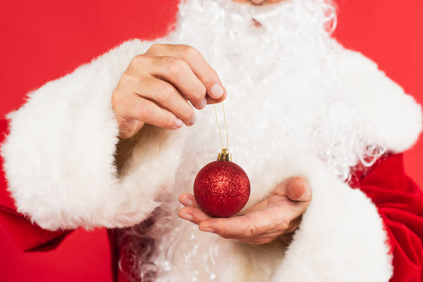 Cropped view of blurred santa holding Christmas ball isolated on red  - Фото, изображение