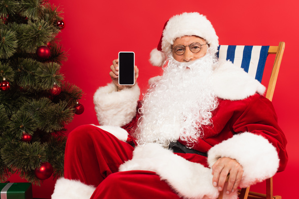 Santa claus holding smartphone on deck chair near christmas tree on red background - Photo, Image
