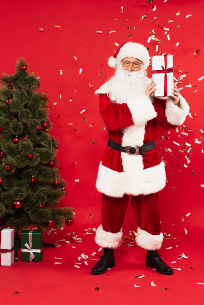 Santa claus holding present near christmas tree and confetti on red background - Photo, image