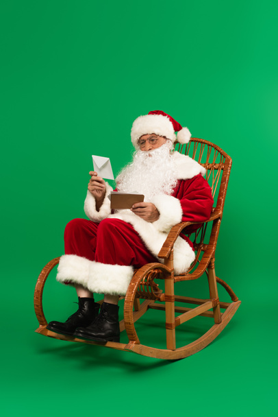Santa Claus sosteniendo sobres en mecedora sobre fondo verde - Foto, Imagen