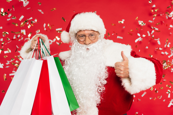 Weihnachtsmann zeigt wie und hält Einkaufstüten unter Konfetti auf rotem Hintergrund - Foto, Bild