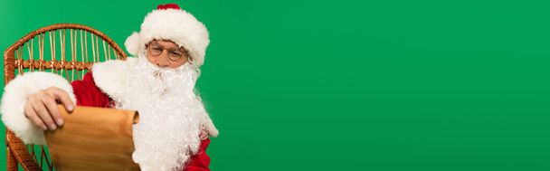 Santa claus holding paper on wicker chair isolated on green, banner  - Foto, imagen