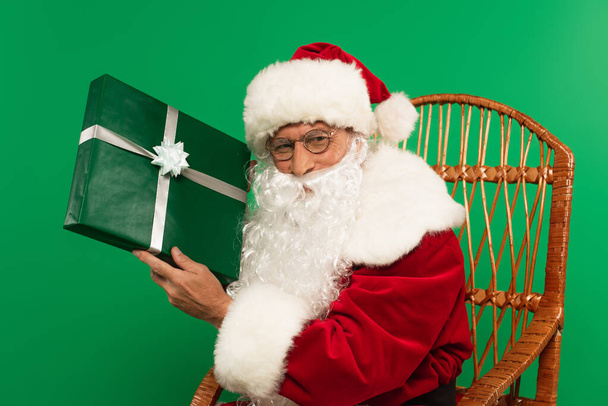 Weihnachtsmann mit Brille hält Geschenk auf Korbstuhl isoliert auf Grün - Foto, Bild