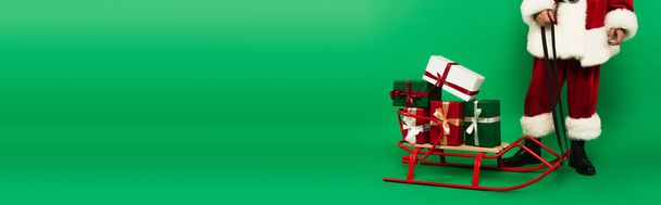 Vista recortada de Santa Claus en traje de pie cerca de regalos en trineo sobre fondo verde, pancarta  - Foto, imagen