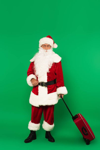 Santa Claus en traje que sostiene la maleta sobre fondo verde - Foto, Imagen