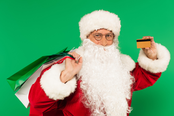 Santa Claus con bolsas de compras y tarjeta de crédito mirando a la cámara aislada en verde - Foto, Imagen