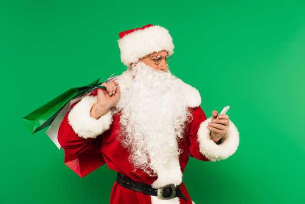 Santa claus with shopping bags using smartphone isolated on green - Фото, зображення