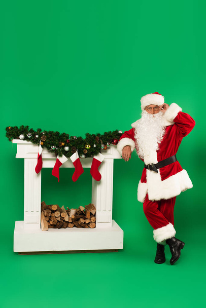 Santa claus adjusting eyeglasses near fireplace with christmas stockings on green background - Photo, Image