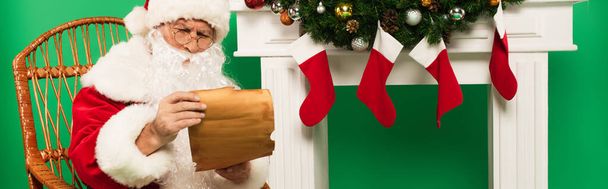 Kerstman houden van papier op schommelstoel in de buurt van open haard met kerst kousen op groene achtergrond, banner  - Foto, afbeelding