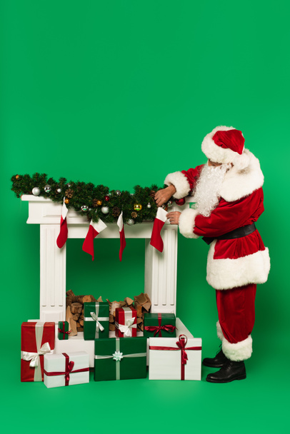 Santa Claude tenant bas de Noël près de cheminée décorée et présente sur fond vert - Photo, image