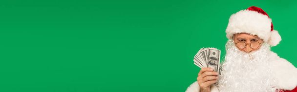 Santa Claus při pohledu do kamery a držení hotovosti izolované na zelené, banner  - Fotografie, Obrázek