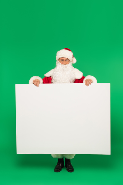 Papai Noel segurando cartaz com espaço de cópia e olhando para a câmera no fundo verde - Foto, Imagem