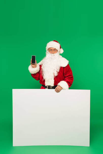 Santa claus holding smartphone with blank screen and placard on green background - 写真・画像