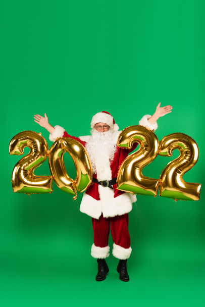 Weihnachtsmann hebt Hände neben Luftballons in Form von 2022 auf grünem Hintergrund - Foto, Bild