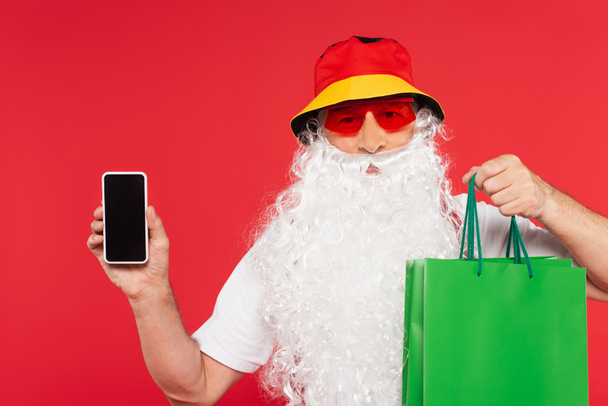 Kerstman in zonnebril met boodschappentassen en smartphone met blanco scherm geïsoleerd op rood  - Foto, afbeelding