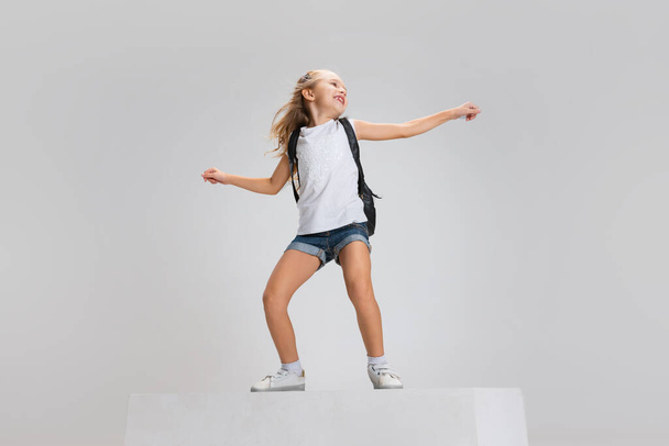 Full-length portrait of smiling girl in casual clothes standing on big box isolated on white studio background. Happy childhood concept. - Photo, image