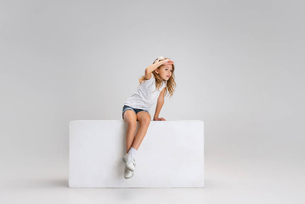 Portrait of little cute girl in casual clothes sitting on big box isolated on white studio background. Happy childhood concept. - Foto, Imagem