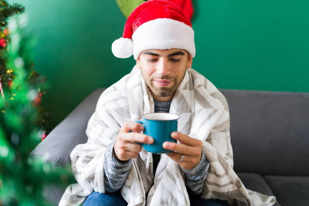 Happy handsome man wrapped in a blanket during a cold Christmas morning while drinking hot chocolate - Photo, Image