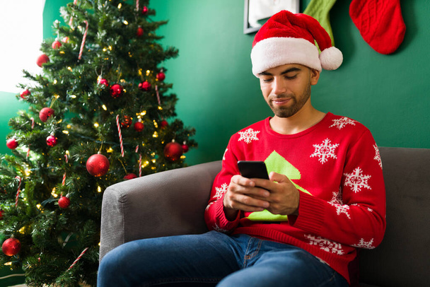 Attraktiver Mann, der auf seinem Smartphone SMS schreibt und soziale Medien nutzt, während er zu Hause Weihnachten feiert  - Foto, Bild