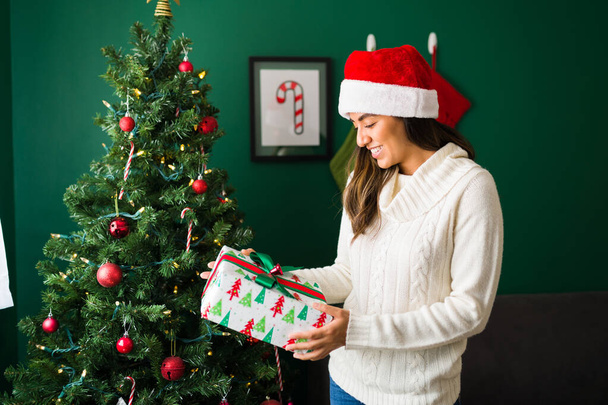 Excited young woman excited about Christmas gifts holding a present next to the tree at home - Foto, Imagen