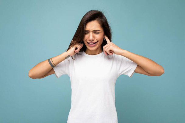 Portrait of angry dissatisfied young beautiful brunette woman with sincere emotions wearing casual white t-shirt for mockup isolated on blue background with empty space and covering ears with fingers - Photo, Image