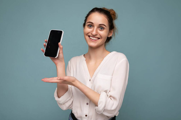 Photo of beautiful smiling young woman good looking wearing casual stylish outfit standing isolated on background with copy space holding smartphone showing phone in hand with empty screen display for - Zdjęcie, obraz