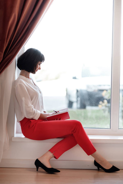 portrait of young European businesswoman with notebook is sitting by the window. Concept of planning. girl in white shirt and red pants - Photo, image