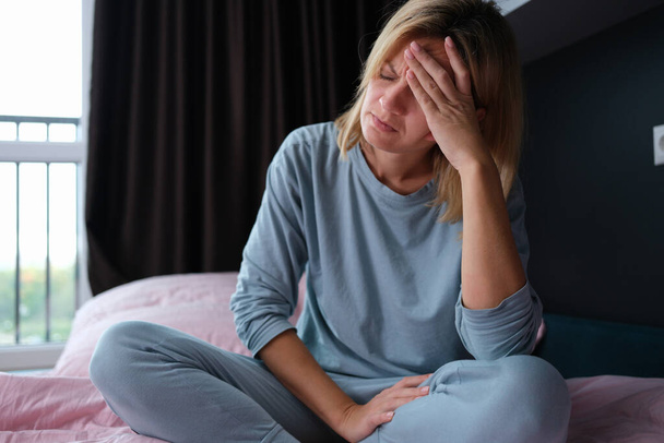 Jeune femme malade en pyjama assise au lit et tenant la tête - Photo, image