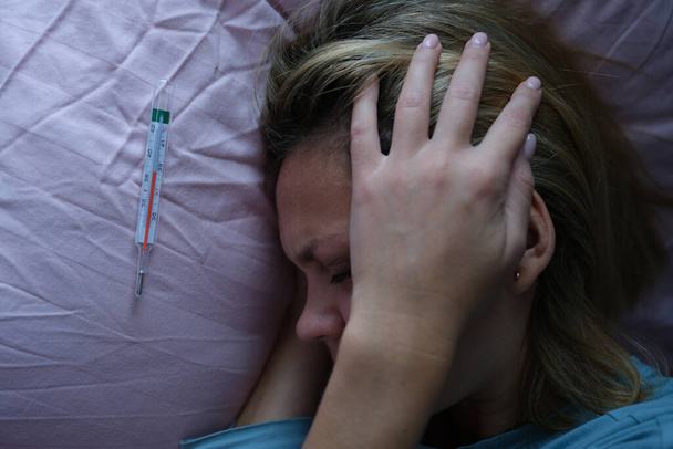 jonge vrouw in bed en houden haar hoofd in de buurt van kwik thermometer - Foto, afbeelding