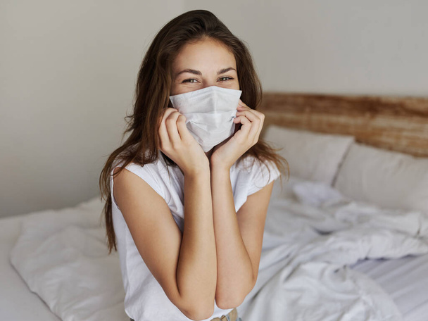 pretty woman in medical mask touches face with hand and sits on bed indoors - Foto, Bild