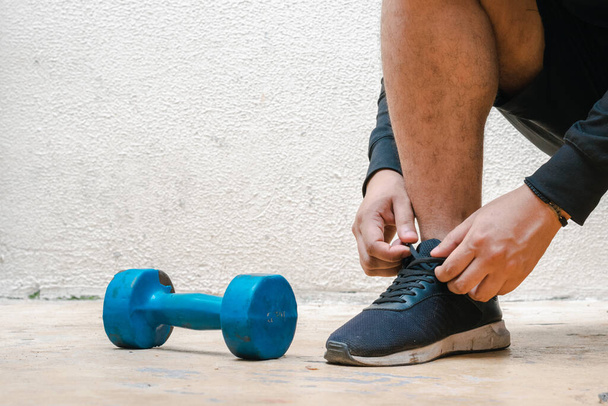 Mann bindet seine Schuhe neben eine blaue Hantel, bevor er Sport treibt. Übung zu Hause, weißer Hintergrund. Fitness-Konzept. Gesundheitskonzept, Platz für Werbetexte - Foto, Bild