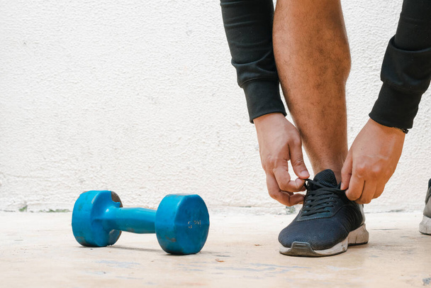 man tying his shoe standing next to blue dumbbell, before exercising. exercise at home, white background. fitness concept. health concept, space for advertising text - Photo, image