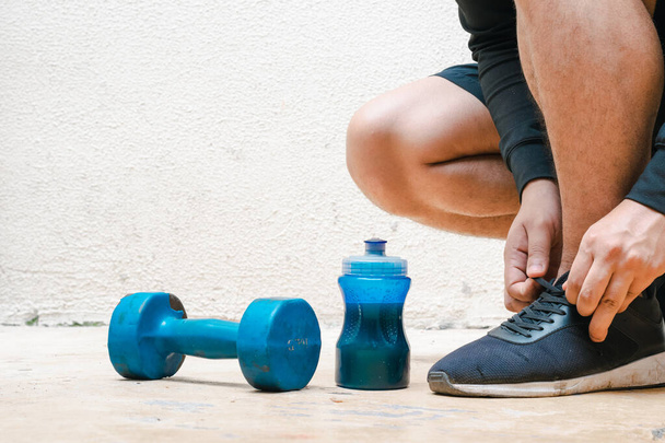man tying his shoes next to dumbbell and bottle with blue water, before exercising. exercise at home, white background. fitness concept. health concept, space for advertising text - Foto, immagini