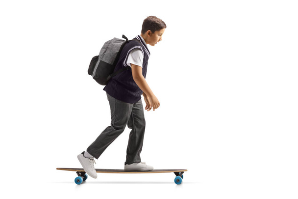 Full length profile shot of a schoolboy in a uniform riding a skateboard isolated on white background - Zdjęcie, obraz