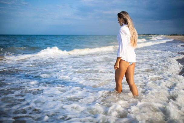 A slender girl in a gentle blue swimsuit and shirt walks along the sandy beach near the blue sea - Фото, зображення