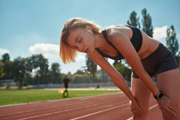 Exhausted sporty young woman taking a break while training, running on the track at stadium on a sunny day - Photo, Image
